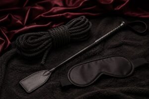BDSM Utensilien wie Peitsche, Seil und Augenbinde auf einer schwarzen Decke